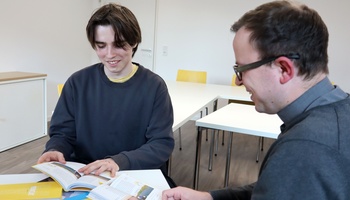 Noah Wirsching und Pfarrvikar Bertram Ziegler reden im Gemeindezentrum St. Andreas in Ochsenfurt. Auf dem Tisch vor ihnen liegen Bücher zur Taufvorbereitung.