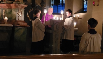 Blick auf den Ambo: Zwei Ministranten stehen rechts und links vom Ambo und halten Leuchter, während der Priester aus der Bibel liest